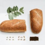 Foto creativa con composizione su sfondo bianco di pane spezzato, rametto con foglie, 6 spine di rosa, 6 sassolini, un rettangolo di terra